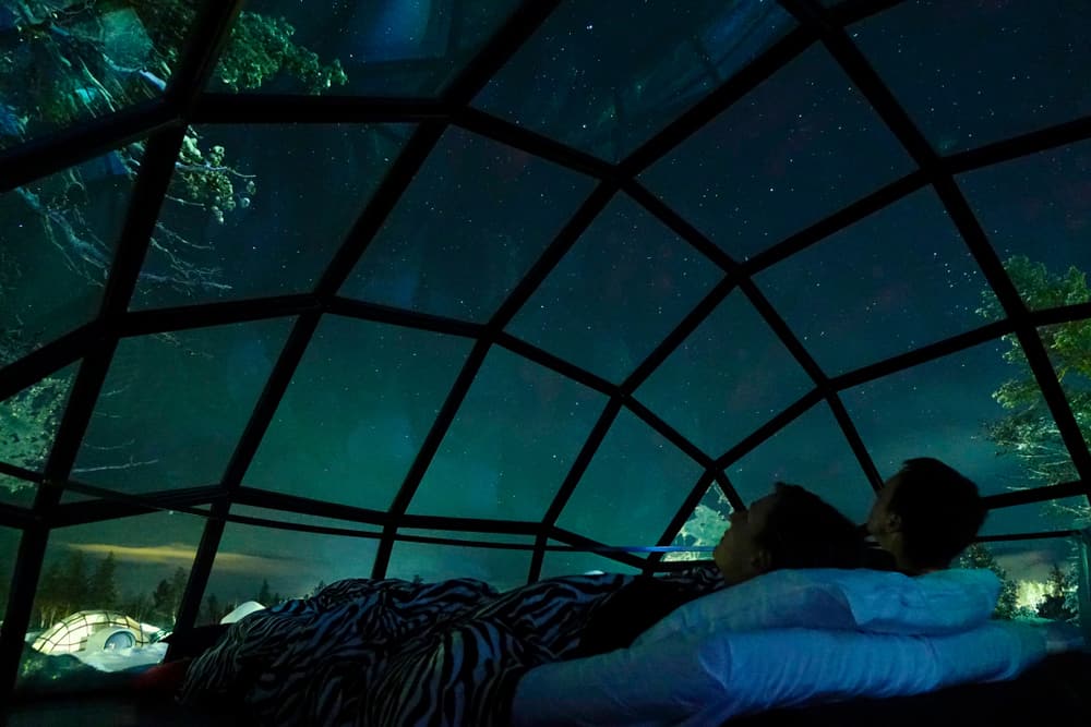 A couple laying in a bed looking up through a glass dome ceiling at the stars 