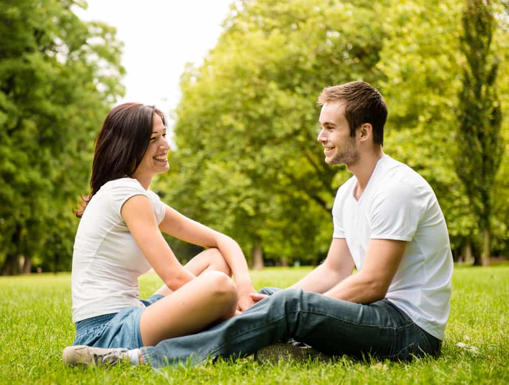 A man and woman are sitting on grass outdoors while smiling.