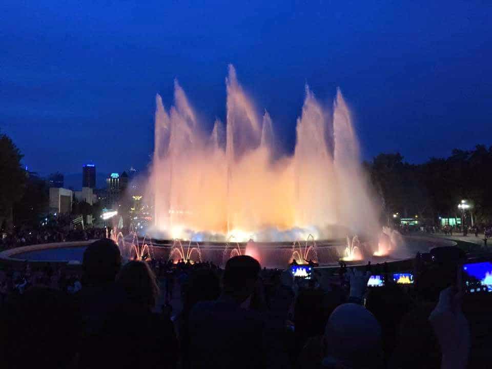 A fountain is lit up at night under a dark sky. People stand near the fountain watching it.